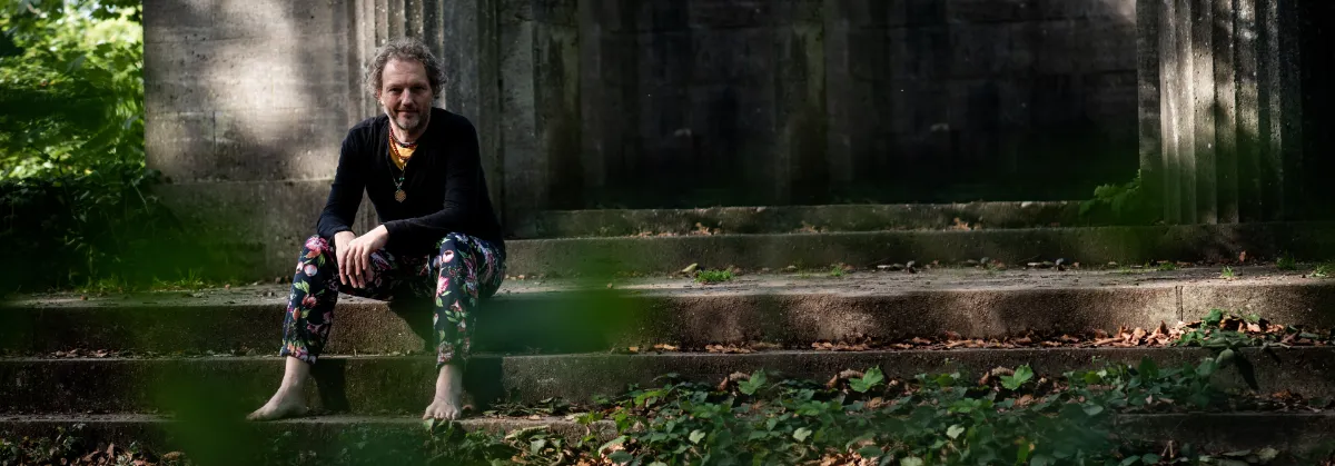 Kink en BDSM coaching: wat is echt waar voor jou? 3 Joris Vincken zit blootsvoets op verweerde betonnen trappen tussen het groen, in een zwarte trui en een broek met bloemenprint
